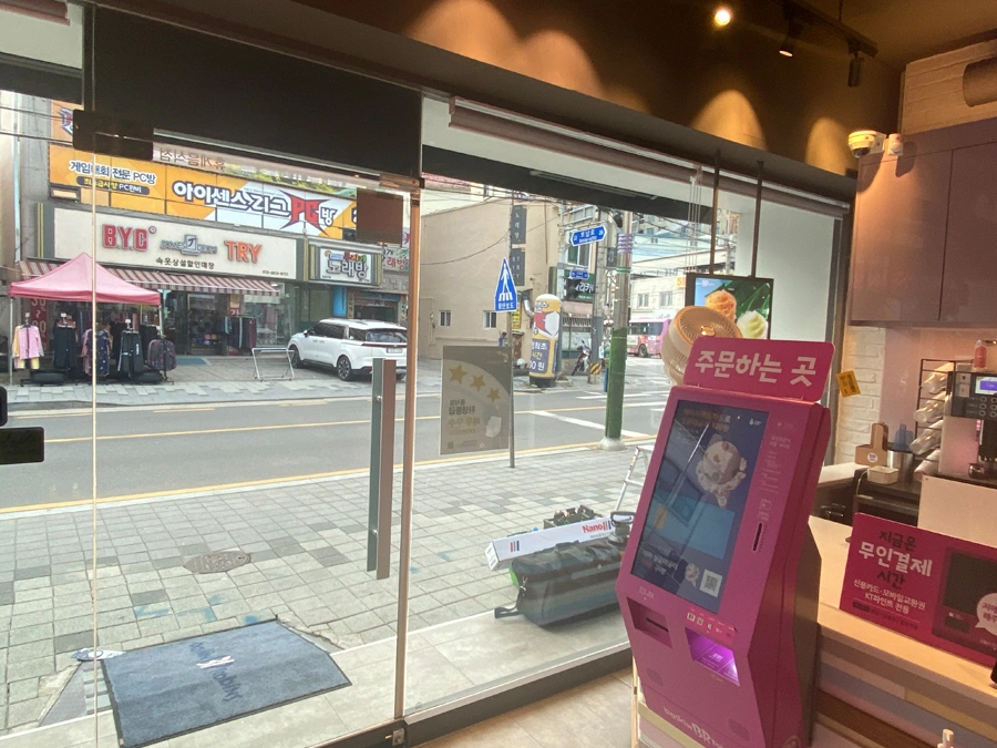 매장 내부 주문 키오스크 옆 통유리창 너머로 맞은편 상가와 도로가 보이는 사진