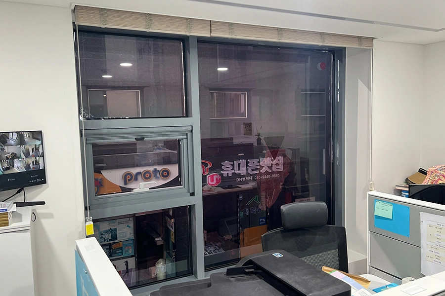 사무실 통유리에 짙은 톤 필름을 시공해 반사가 강해진 창문과 책상, CCTV 모니터가 보이는 사진