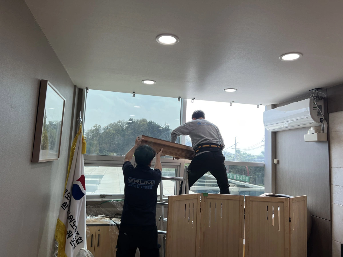 태극기가 걸린 사무 공간 창가에서 두 명의 작업자가 긴 필름을 함께 들어 옮기는 사진